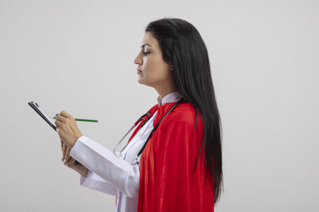 穿着专注的年轻白种人超级英雄女孩戴着听诊器站在侧视图上用铅笔在剪贴板上写字 在白色背景上隔离 留有复印空间复制超级英雄集中
