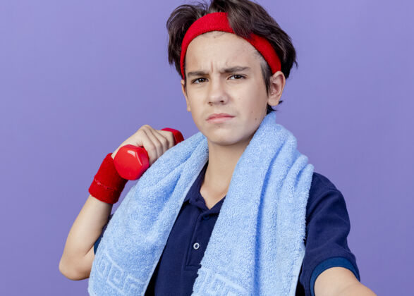 运动特写镜头 年轻帅气的运动男孩戴着头带和护腕 戴着牙套 脖子上围着毛巾 举着哑铃 看着紫色墙壁上孤立的前方表情毛巾不愉快