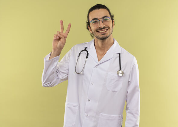 微笑微笑的年轻男医生戴着眼镜 身穿白色长袍 手持听诊器 在绿色上显示和平姿态和平空间男性