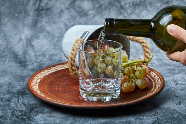 深色小桶内的葡萄陶瓷盘子和手倒入大理石背景上的玻璃酒果汁盘子食物