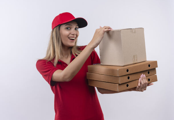 欢乐快乐的年轻送货女孩穿着红色制服和帽子举行许多箱子隔离在白色的墙壁上穿帽子盒子