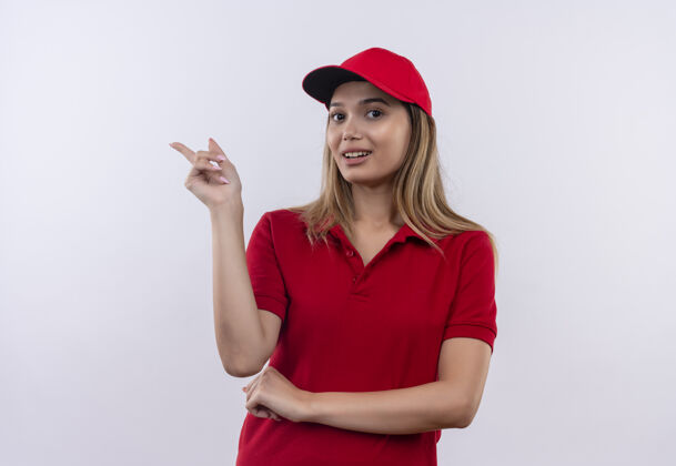 壁板高兴的年轻送货女孩穿着红色制服 戴着帽子指向侧面 隔离在白色墙上 留有复印空间送货请穿制服