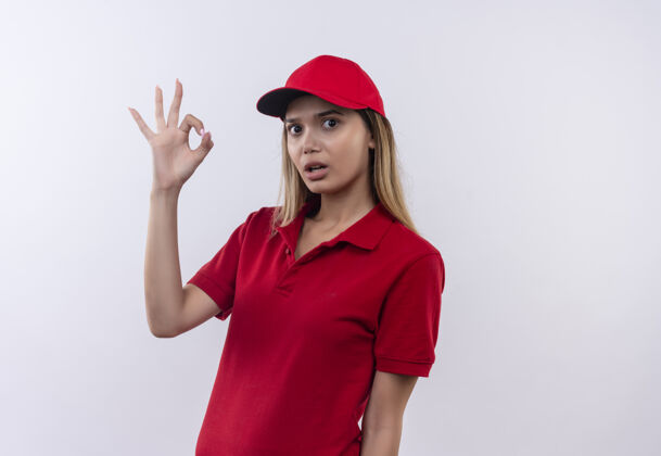 穿一个穿着红色制服 戴着帽子的年轻送货女孩惊讶地站在白色的墙上 表现出很好的姿势好的展示帽子
