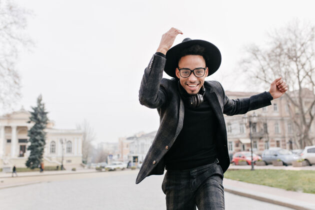 公园一个戴眼镜的非洲男人在秋天的公园里奔跑一个戴着帽子的时髦黑人在城市广场上嬉戏的户外肖像优雅城镇放松
