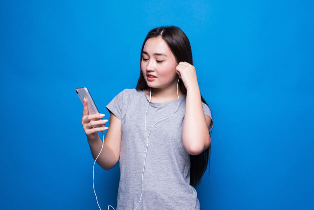 人年轻的亚洲女人在蓝色的无缝墙里用红色耳机听音乐娱乐 音乐应用 网上热气腾腾音乐年轻电话
