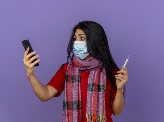 人心急如焚的年轻病妇戴着口罩 戴着围巾 拿着手机和体温计 看着隔离在紫色墙上的手机感觉电话公民