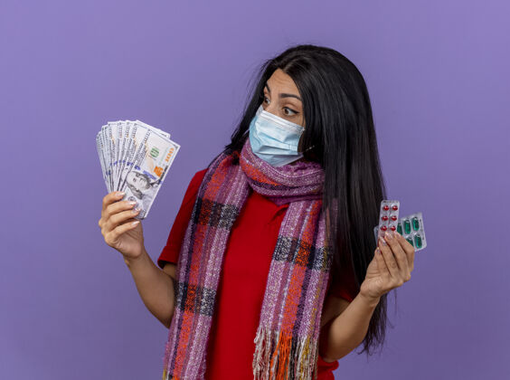 人印象深刻的年轻生病的女人戴着面具和围巾拿着钱和一包胶囊看着隔离在紫色墙上的钱站围巾钱