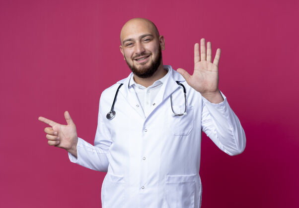墙高兴的年轻秃头男医生穿着医用长袍和听诊器显示不同的数字隔离在粉红色的墙上穿长袍医生