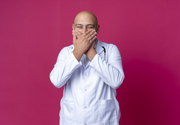 年轻年轻的秃顶男医生穿着医用长袍 戴着听诊器 笑着用手捂住嘴巴 隔离在粉红色的墙上罩子医生听诊器