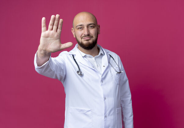 穿高兴的年轻秃头男医生穿着医用长袍和听诊器显示停止手势隔离在粉红色的墙上请年轻停止