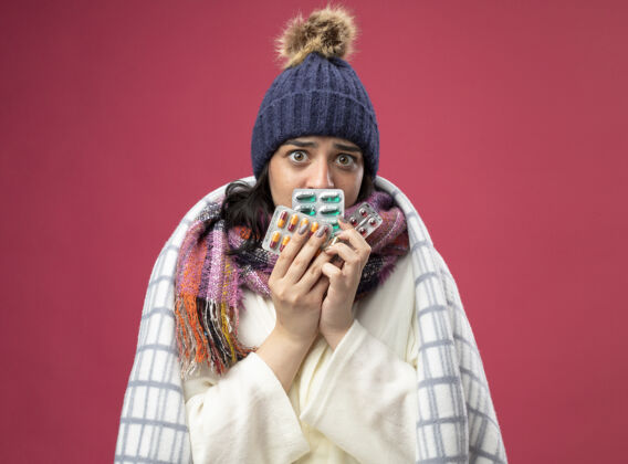 女人印象深刻的年轻生病的女人穿着长袍 戴着冬天的帽子和围巾裹在格子布手里拿着一包包胶囊在前面的嘴上看着前面孤立的粉色墙壁脸人穿