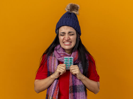 围巾恼怒的年轻生病的女人戴着冬天的帽子和围巾拿着一包胶囊和其他包在帽子下面闭着眼睛隔离在橙色的墙上关冬天眼睛