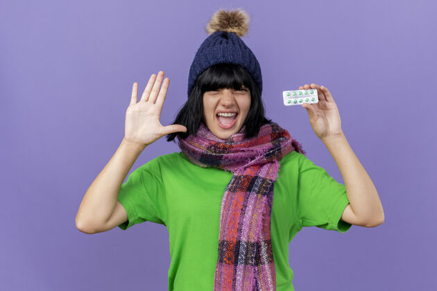 脸自信的生病的年轻女人戴着冬天的帽子和围巾 看着前面拿着一包胶囊 在紫色的墙上显示出孤立的空手穿空感情
