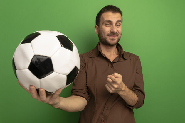 姿势高兴的年轻人把足球伸到前面 看着摄像机指着隔离在绿墙上的球公民站立人
