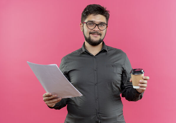 咖啡高兴的年轻商人戴着眼镜拿着一杯咖啡和纸隔离在粉红色的墙上请穿上杯子年轻人