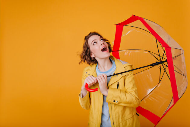 雨一个穿着时髦外套的无忧无虑的女孩拿着雨伞 抬头看了看黄色墙上一个卷发美女和阳伞合影的摄影棚镜头雨季节漂亮