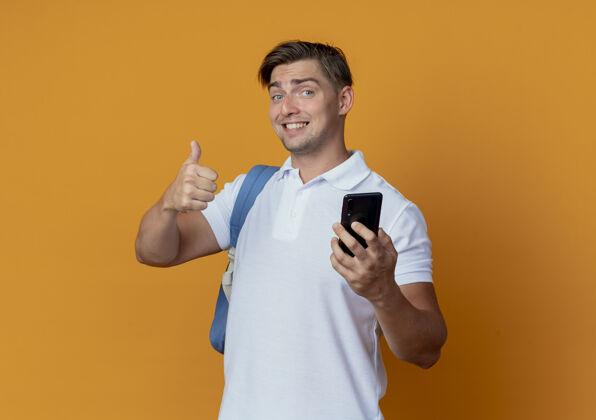 向上微笑着的年轻帅气的男学生背着书包拿着手机大拇指孤立地竖起放在橘子上包拇指年轻