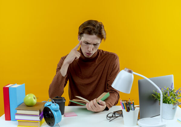 背景年轻的学生男孩坐在书桌旁 手里拿着学习工具 手放在黄色的下巴上看书工具坐着思考