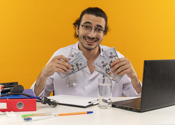 笔记本电脑微笑的年轻男医生戴着医用眼镜 穿着带听诊器的医用长袍工作听诊器长袍