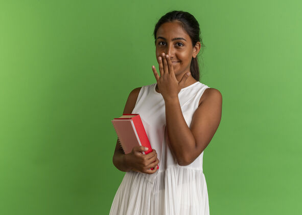 手高兴的年轻女学生拿着笔记本 用手捂着嘴放在绿色的纸上请盖上嘴女学生