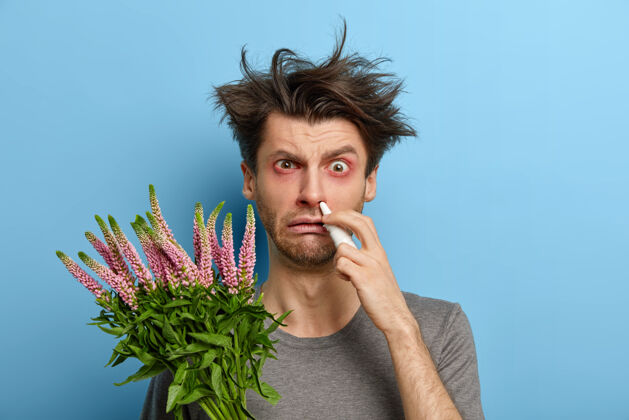 医学过敏的男人发型凌乱 眼睛发红发痒 拿着植物打喷嚏或僵硬 有不愉快的症状 在家治疗 靠着蓝墙站着鼻涕脸问题肿胀