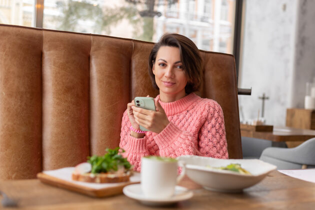 女孩餐厅里一位穿着暖和毛衣的女士正在看电话里的东西 桌上摆着美味健康的早餐和抹茶拿铁肖像美味人