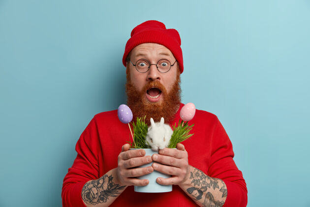 春天一个留着胡子的时髦小伙拿着一个罐子 里面放着白色蓬松的复活节小兔子和装饰过的彩蛋 象征着春天和节日 戴着红帽子 套头衫和眼镜 在蓝色的墙上摆姿势狡猾持有凝视
