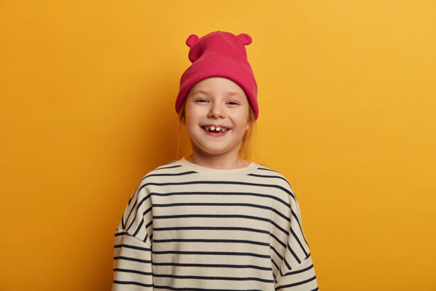 毛衣可爱的小孩子咯咯笑着积极地缺牙 心情愉快 欢庆美好的一天 戴着粉色时髦的帽子和条纹宽松的毛衣 对着黄色的墙壁摆姿势 拍下难忘的照片感觉情绪快乐