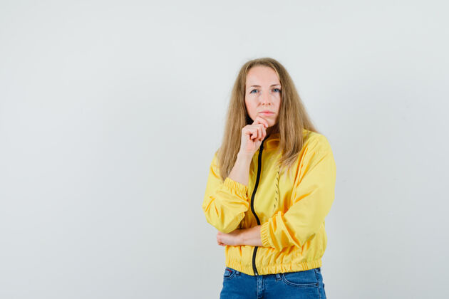 女性身着黄色棒球服和蓝色牛仔裤的年轻女子站在思考的姿势中 面带沉思 俯视前方夹克姿势年轻