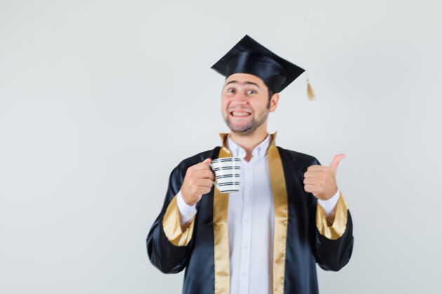 男人年轻人手里拿着一杯咖啡 穿着毕业制服竖起大拇指 看上去很快乐 正对着前方拇指年轻毕业