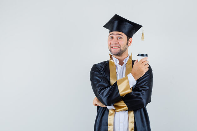 杯子年轻的男性拿着一杯咖啡 穿着研究生制服 看上去很高兴正面图模特衬衫毕业