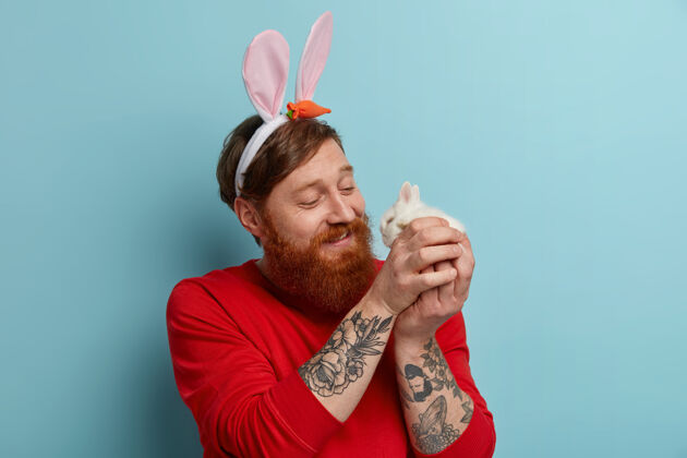 兔子关心快乐的姜胡子男人和可爱的小兔子玩耍 戴着兔子耳朵和红色毛衣 庆祝复活节 享受春天 在室内摆姿势传统和宗教节日的概念满意胡须欧洲