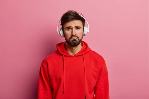 人失望沮丧不快乐的男人试图用音乐来娱乐自己 忧郁的表情 戴着耳机 穿着红色连帽衫 隔离在玫瑰色的粉彩墙上胡须忧郁音乐