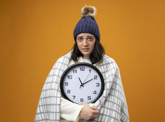 表情虚弱的生病的年轻女人穿着长袍 戴着冬天的帽子 裹着格子布 拿着钟 看着橙色的墙上孤立的前方女人衣服站着
