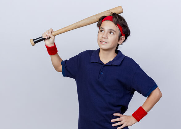 人体贴的年轻帅气的运动男孩 戴着头带和带牙套的腕带 手放在腰上 抬起头 用隔离在白墙上的棒球棒碰着头站立背带保持