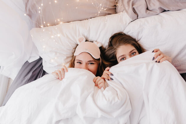 漂亮漂亮的白人女孩躲在白色的毯子下令人惊叹的年轻姐妹在早晨摆姿势的室内画像床睡衣微笑