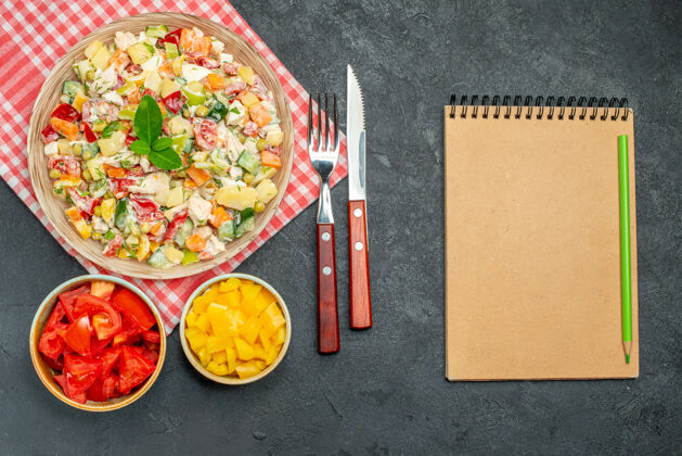 学校一碗蔬菜沙拉的顶视图 红色餐巾上有蔬菜餐具和记事本 放在深色的桌子上桌子蜡笔纸
