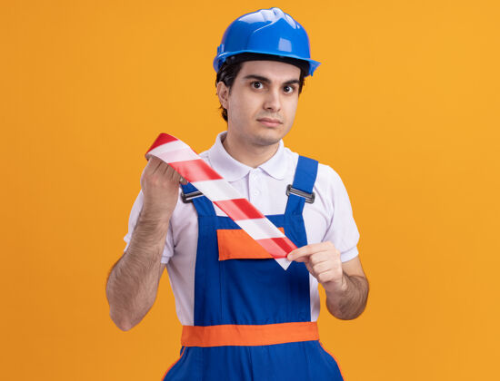 表情年轻的建筑工人穿着建筑制服 戴着安全帽 拿着警示带 带着严肃自信的表情站在橙色的墙上 看着前方安全抱着制服