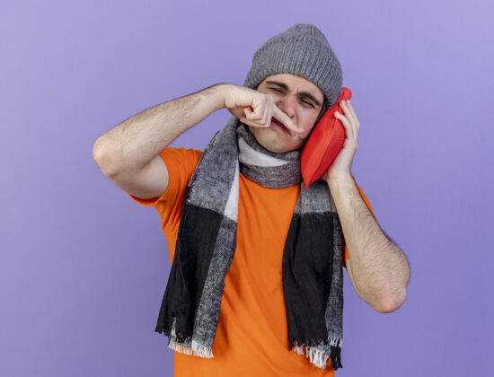 包闭着眼睛 戴着带围巾的冬帽的年轻病人 把热水袋放在脸颊上 用手在紫色背景上擦鼻子手脸颊眼睛