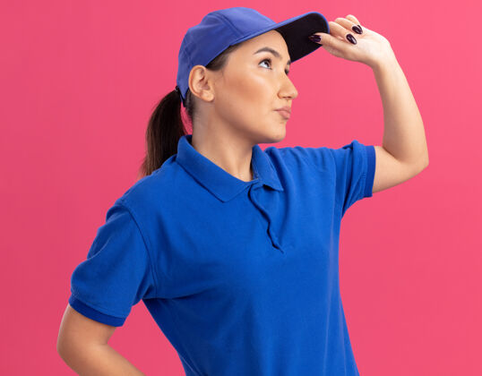 站立身穿蓝色制服 戴着帽子的年轻送货员站在粉红色的墙上 茫然地向一旁望去帽子制服年轻