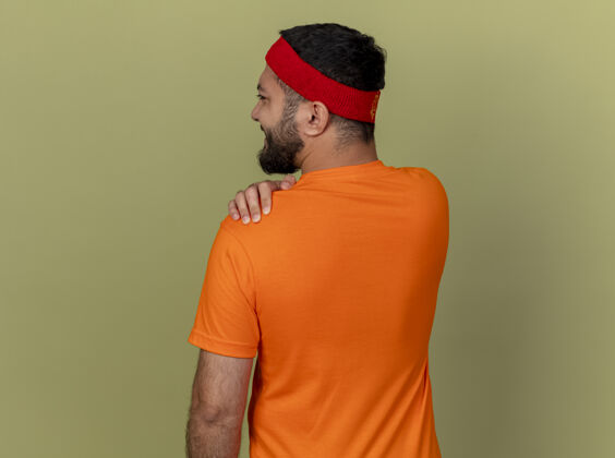 站立背对着镜头站着 戴着头带和腕带的年轻运动型男子抓住了橄榄绿背景上疼痛的肩膀相机年轻肩膀