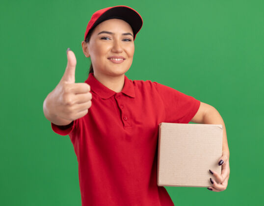 年轻身着红色制服 头戴鸭舌帽 拿着纸板箱 面带微笑 高高兴兴地竖起大拇指站在绿色墙壁上的快乐年轻送货员拇指微笑表演