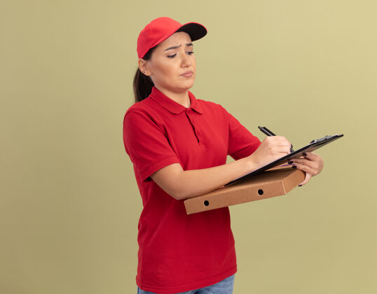年轻人身穿红色制服 头戴鸭舌帽的年轻送货员站在绿色的墙上 手里拿着披萨盒 剪贴板和铅笔 表情严肃脸拿着女人
