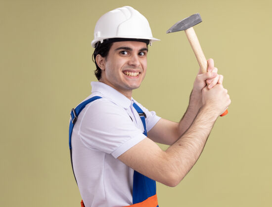 建筑年轻的建筑工人身穿建筑制服 戴着安全帽 手持铁锤 站在绿色的墙上 狡猾地微笑着看着前方站着男人头盔
