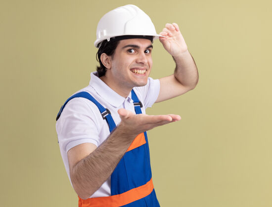 欢呼年轻的建筑工人身穿建筑制服 戴着安全帽 伸出手臂 站在绿色的墙壁上 面带微笑 望着前方制服手臂头盔