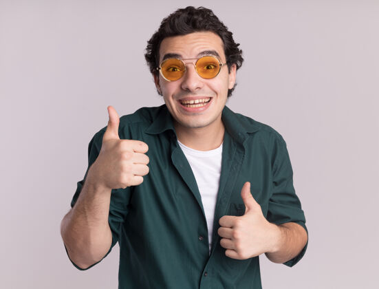 微笑身穿绿衬衫 戴着眼镜的年轻人站在白色的墙上 开心地 积极地微笑着 竖起大拇指眼镜年轻展示
