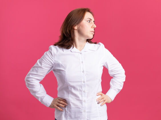 衬衫穿白衬衫的年轻女子站在粉红色的墙上 面带严肃的神情 双臂放在臀部严肃女人手臂