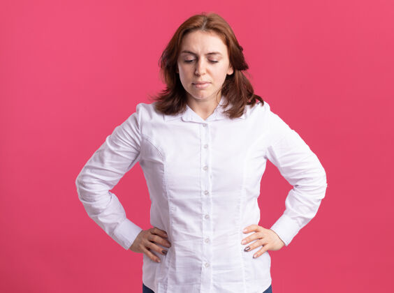 手臂身穿白衬衫的年轻女子 面带严肃的表情 双臂放在臀部 站在粉红色的墙上站立女人严肃