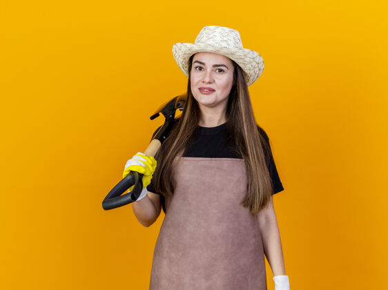 制服微笑美丽的园丁女孩穿着制服和园艺手套帽子把铁锹放在肩膀上孤立的橙色背景女孩肩膀黑桃