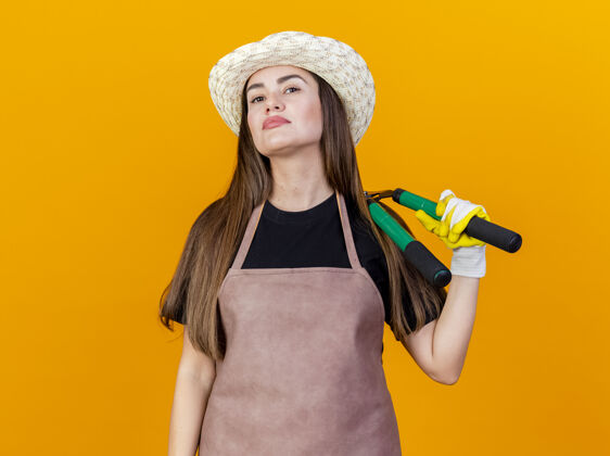 肩膀自信美丽的园丁女孩穿着制服 戴着园艺帽 戴着手套 肩上拿着剪子 在橙色背景上隔离开来手握手套自信